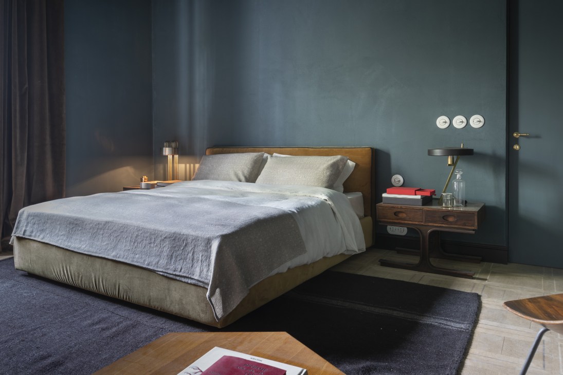 Modern bedroom at Sister Hotel with bed, wooden nightstand, and blue-grey wall with porcelain light switches.