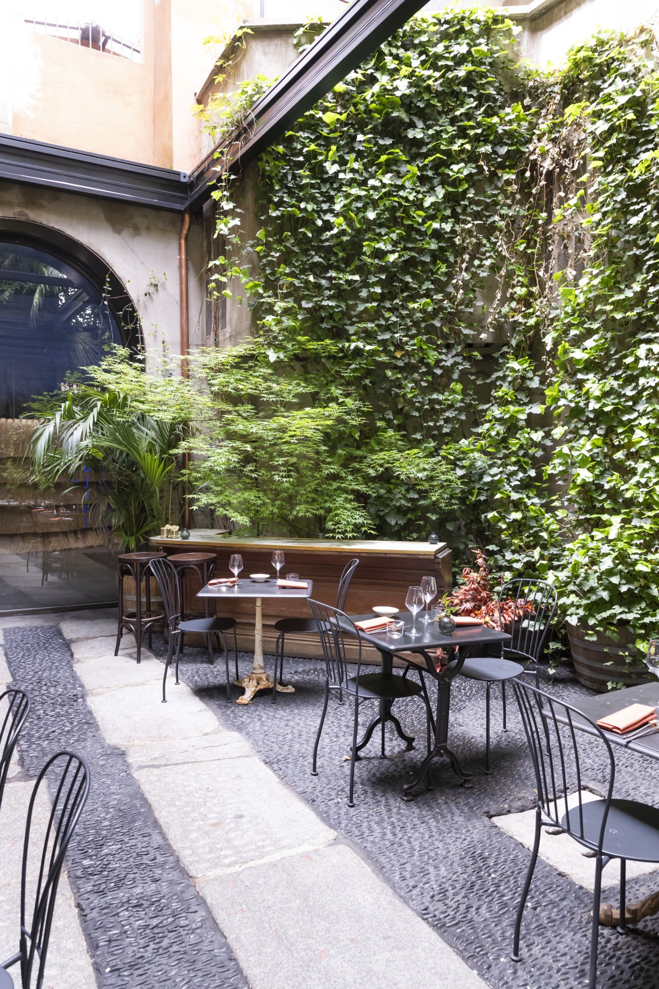 Ivy-covered courtyard at Six in Milan with dining tables and black chairs arranged on patterned flooring.