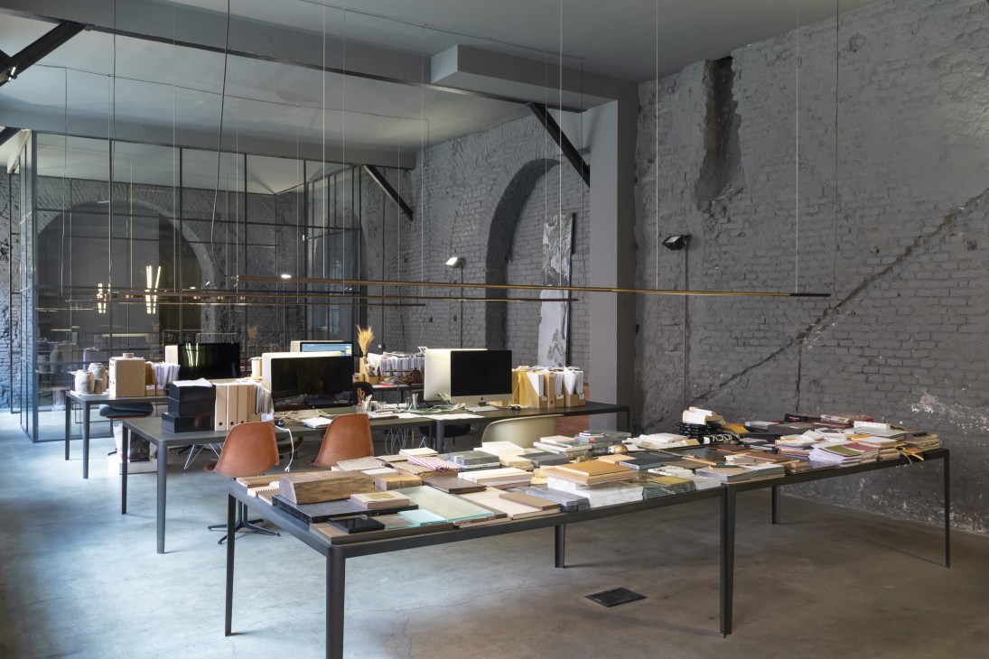 Industrial workspace with exposed brick walls, glass partitions, wooden desks, computer monitors, and pendant lighting in a renovated building.