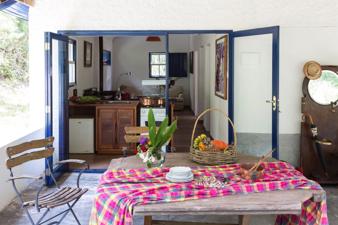 Outdoor dining area with blue-framed doors opening to a kitchen, colorful patterned tablecloth, and fresh produce basket at Casas de Catuçaba.