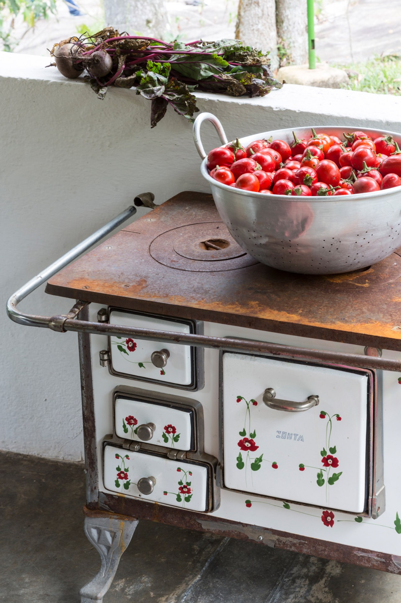 Fresh red tomatoes in a metal colander next to beets on a rustic stove with floral decorative details.