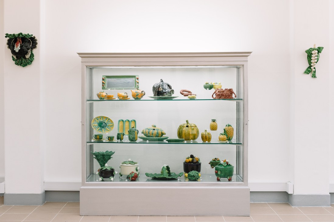 Display cabinet showing Bordallo Pinheiro ceramic pieces including cabbage-shaped bowls, fruit tureens, and vegetable-form vessels in greens and yellows.