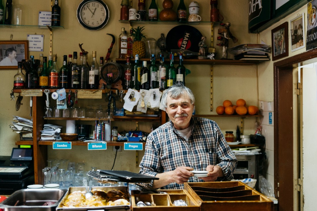 Abilio José Miguel, owner of Cantinho do Alfredo, sits smiling behind the counter of his traditional Portuguese tasca