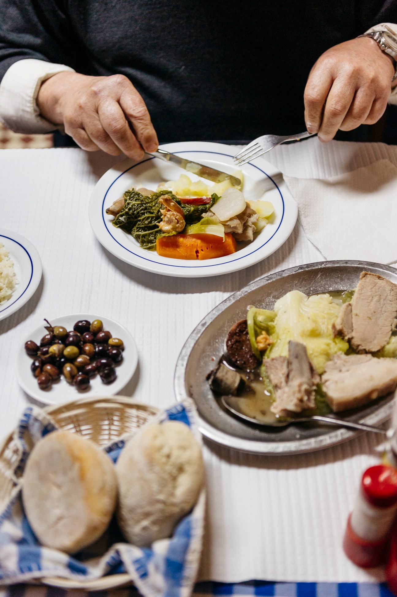 Person eating traditional Portuguese meal with vegetables, olives, bread and soup at white table
