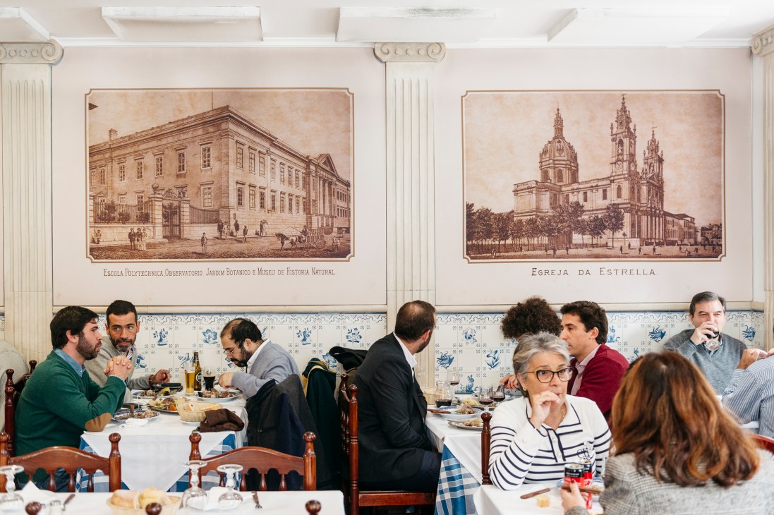Diners eating at tables in Casa dos Pasarinhos restaurant with historic Lisbon paintings on white tiled walls