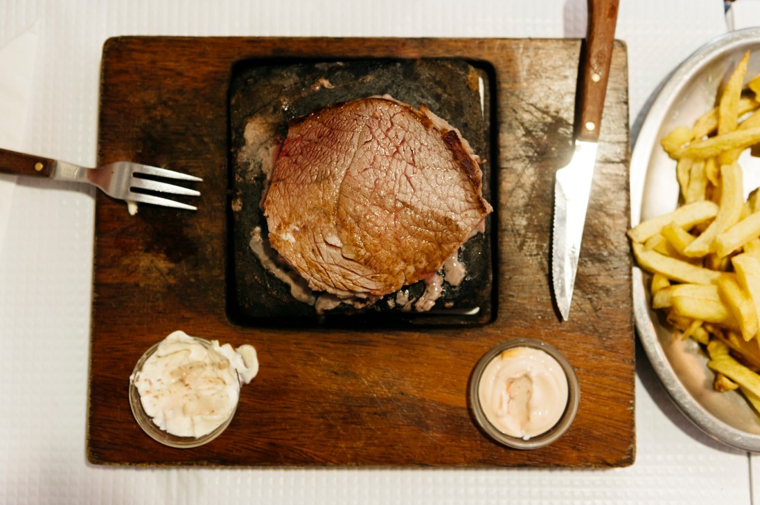 Sliced meat served on black stone plate with two small bowls of sauce and side of french fries