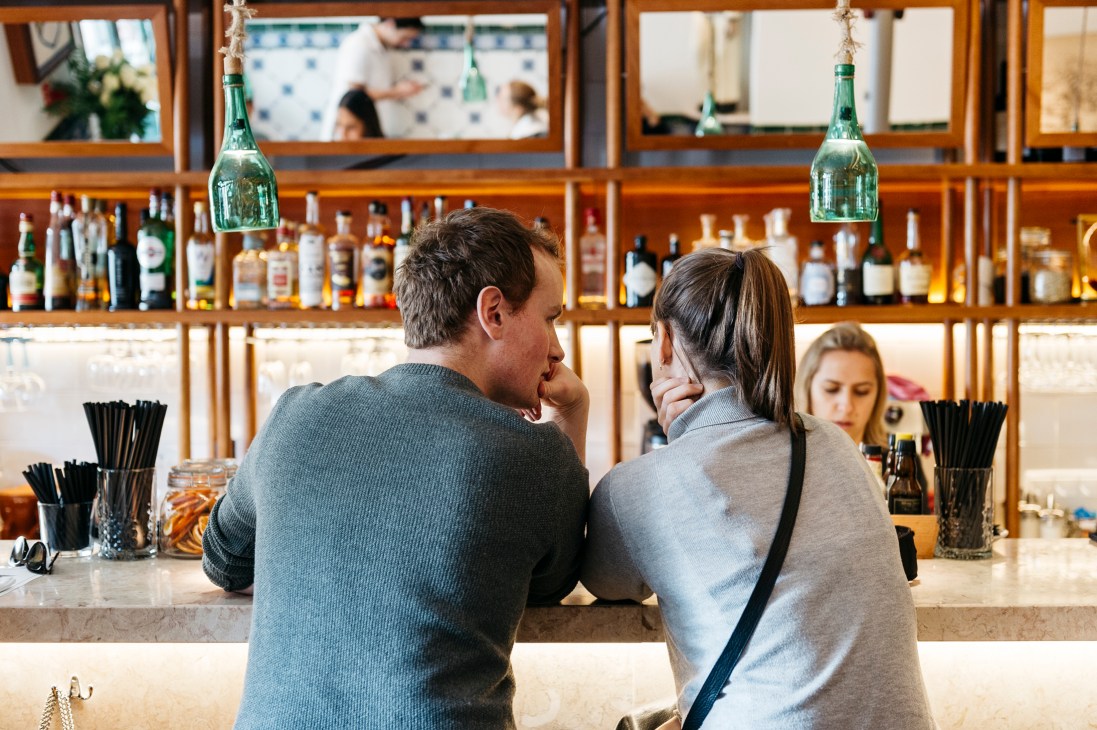 Two people sitting at a bar counter in a traditional Portuguese tasca restaurant with bottles and glasses visible