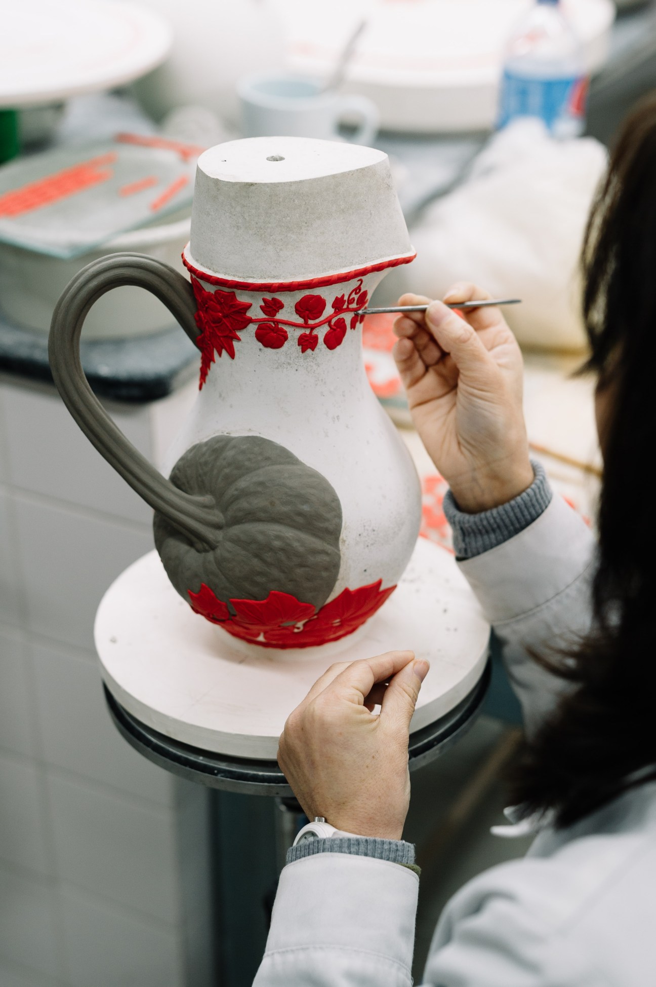A craftsperson hand-paints red floral details onto a white ceramic pitcher with a sculptural handle in the studio.
