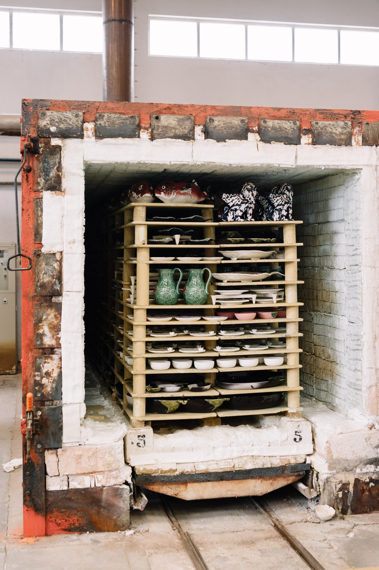 Ceramic pieces arranged on wooden shelves inside an industrial kiln with a red metal frame and white interior.