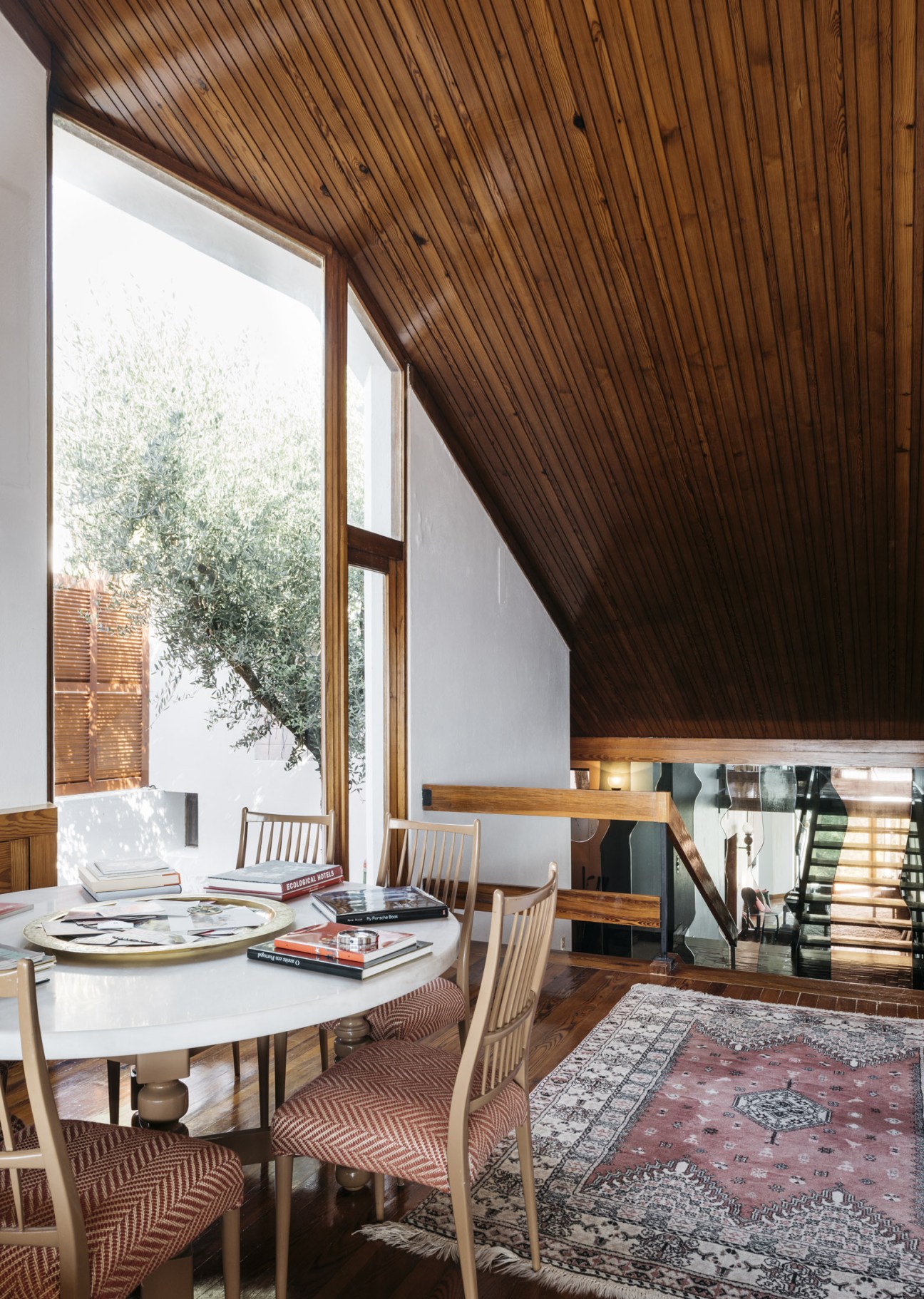 Modernist dining area with wooden slatted ceiling, round white table, and staircase with glass balustrade.