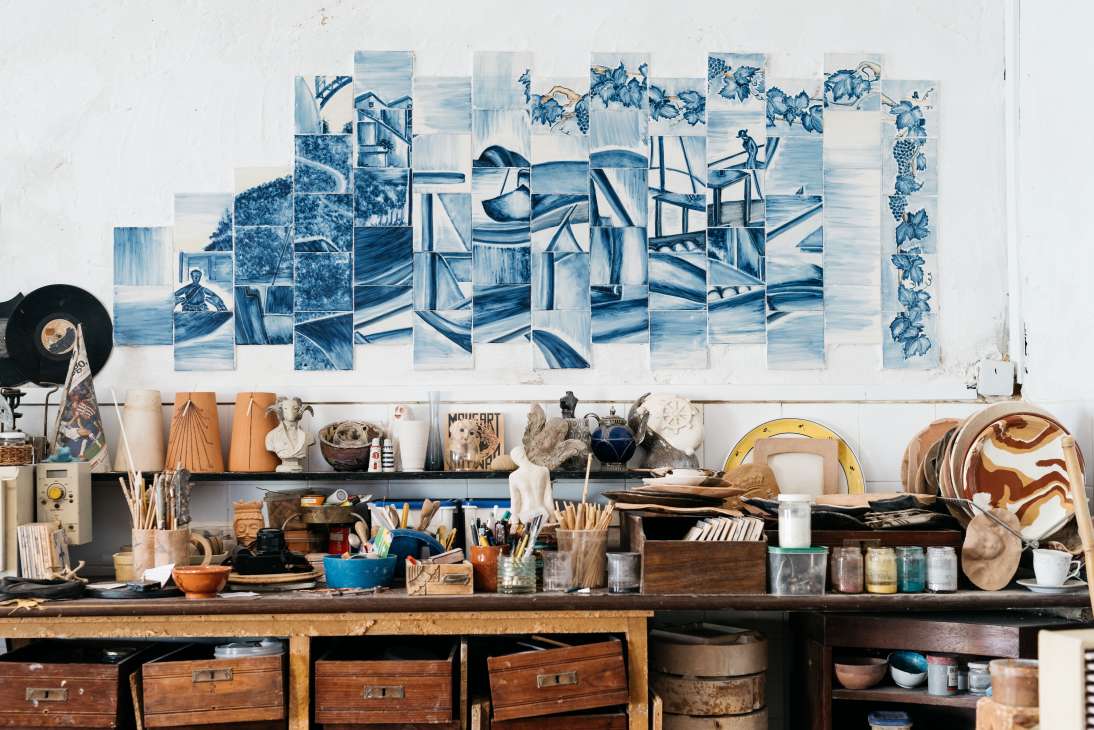 Célia Mendes's ceramic workshop with blue and white tiles arranged on a white wall above a cluttered worktable.