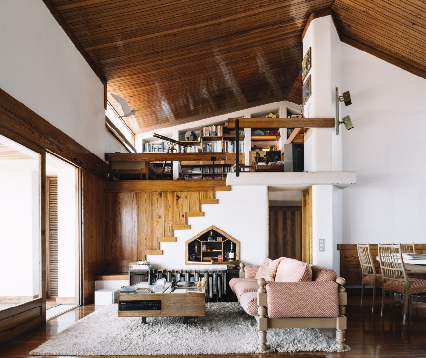Modernist living room with varnished wood ceiling, white walls, wooden staircase, pink armchair and built-in shelving.