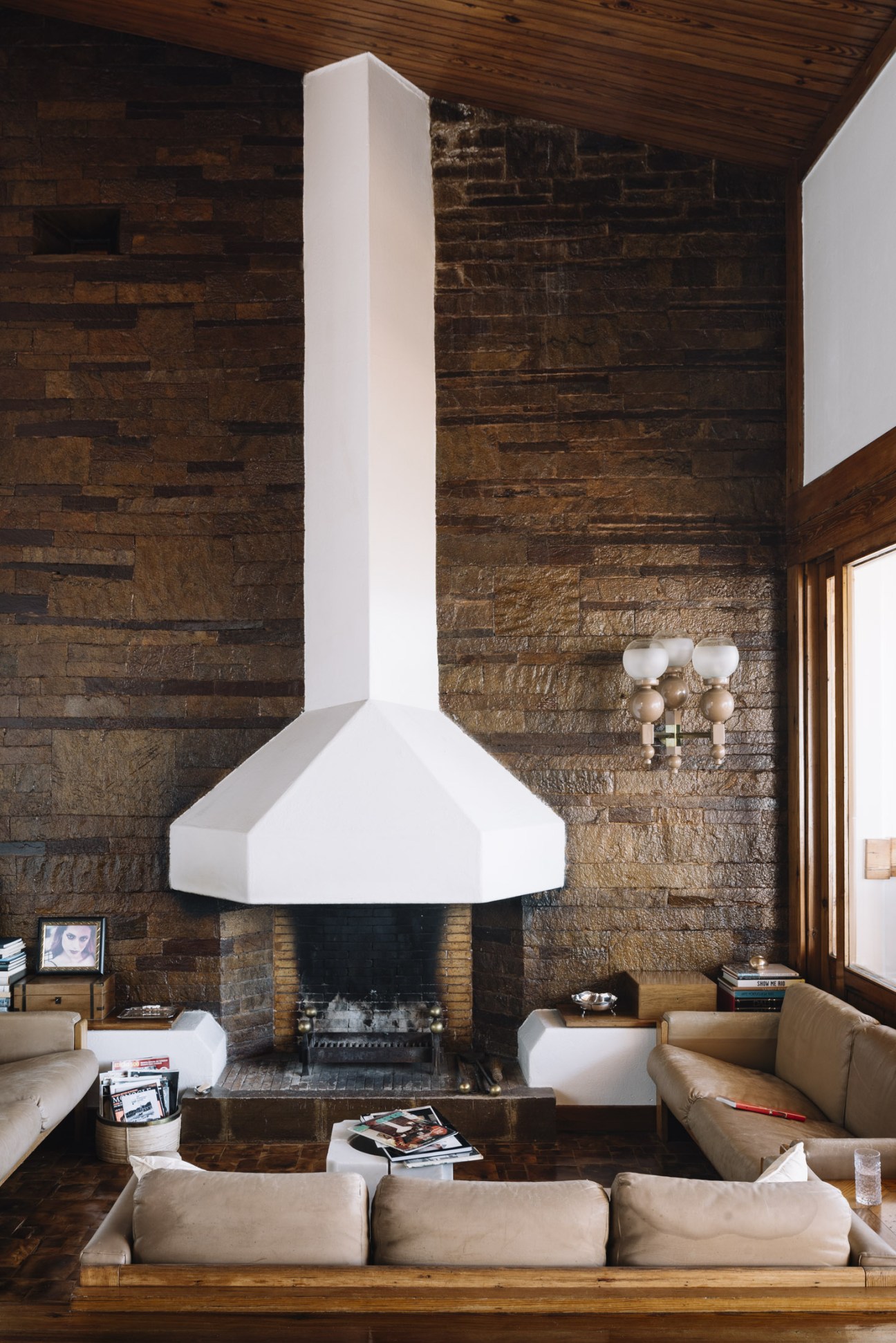 Modernist fireplace with white hood against Portuguese limestone wall in living room with tan seating.