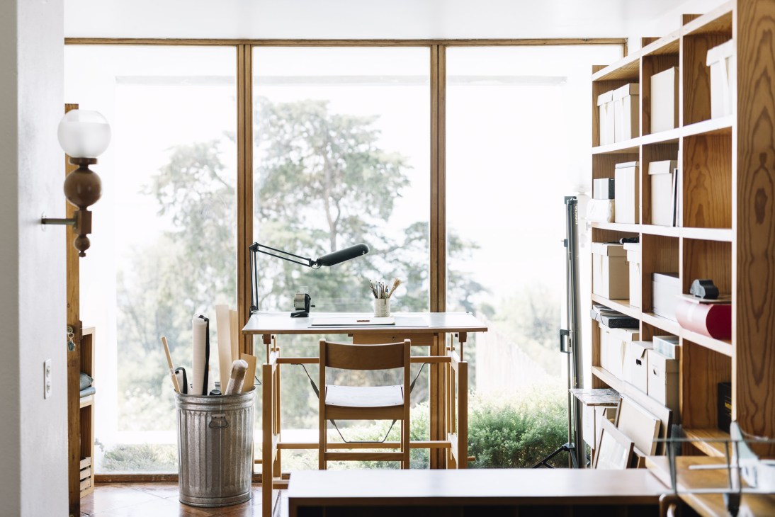 Study nook with wooden desk, chair, desk lamp, and built-in shelving overlooking garden through large windows.