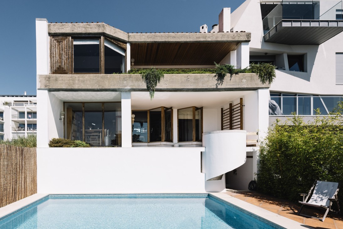 Modernist house with white walls, concrete terraces, and wooden elements overlooks a swimming pool under clear blue sky.