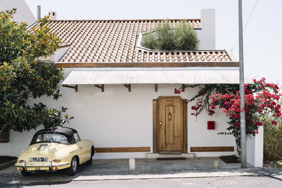 Cream-coloured vintage Porsche parked in front of a modernist white-walled house with terracotta tiles and wooden door.