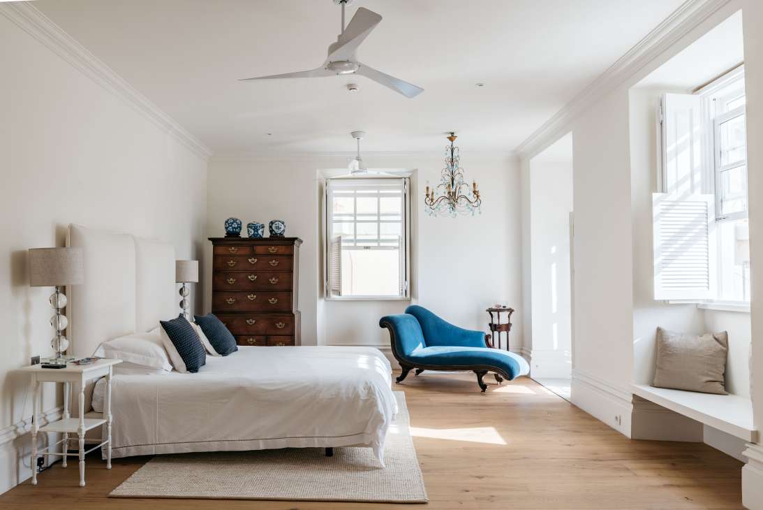 Bright bedroom with white bed, blue chaise lounge, wooden chest of drawers, and chandelier at Casa Fuzetta.