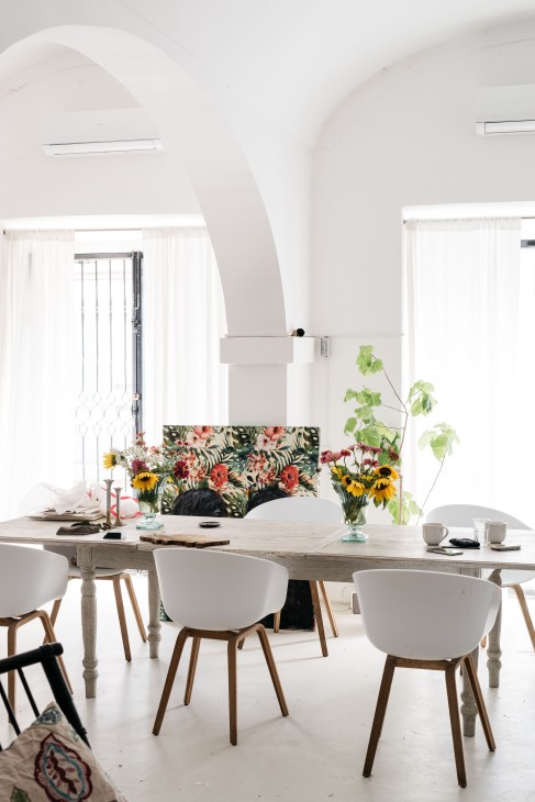Modern dining room with arched white ceiling, wooden table, white chairs with wooden legs, and colorful floral arrangements in glass vases.