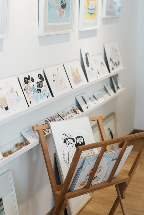 White shelves displaying framed illustrations and prints, with a wooden stand holding art portfolios and sketchbooks in the foreground.