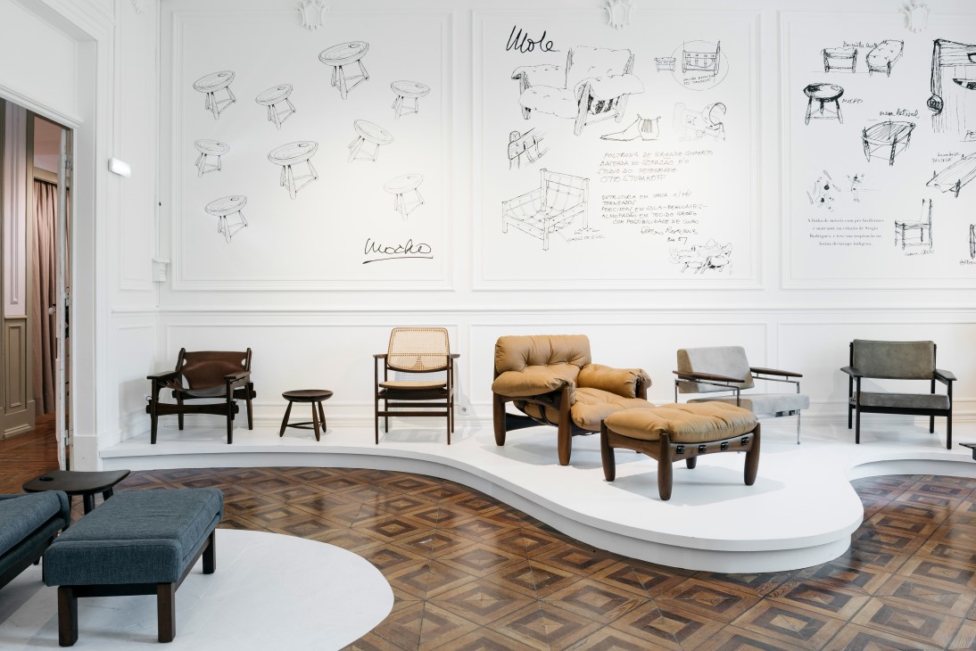 Modernist chairs displayed on white platforms, including tan leather and wood pieces by Brazilian designer Sérgio Rodrigues, with design sketches on the wall above.