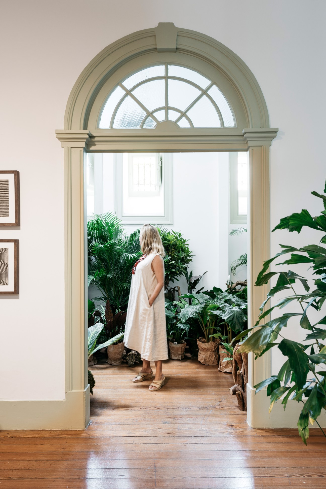 Woman entering a bright room filled with tropical plants through a classical arched doorway with transom window.