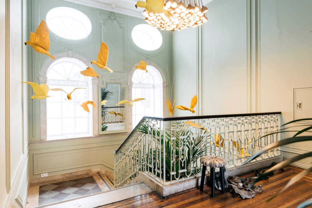 Restored staircase in Palácio Castilho with yellow bird sculptures hanging from ceiling and tropical plants.