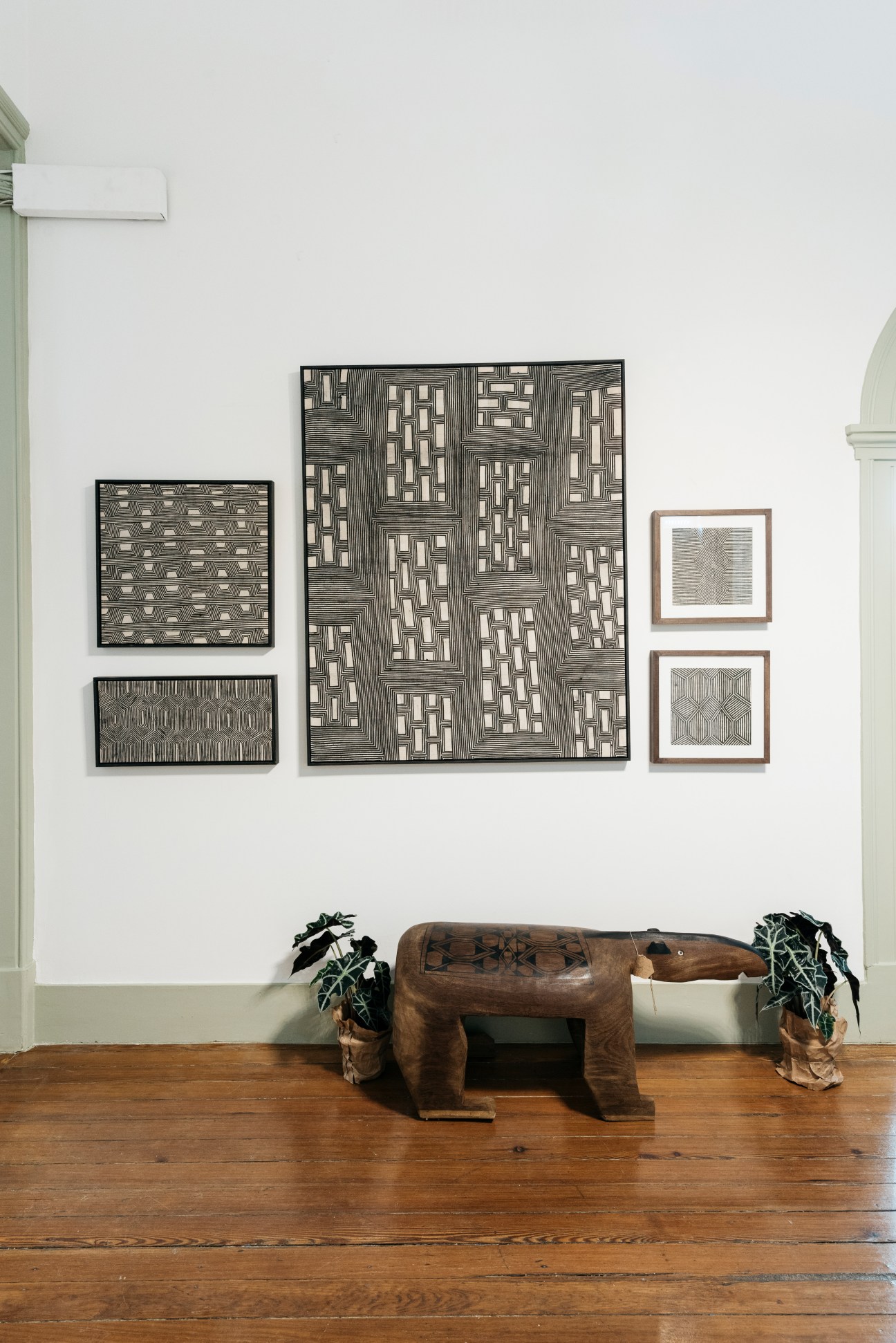 Framed geometric prints displayed on a white wall above a wooden carved animal sculpture with potted plants.