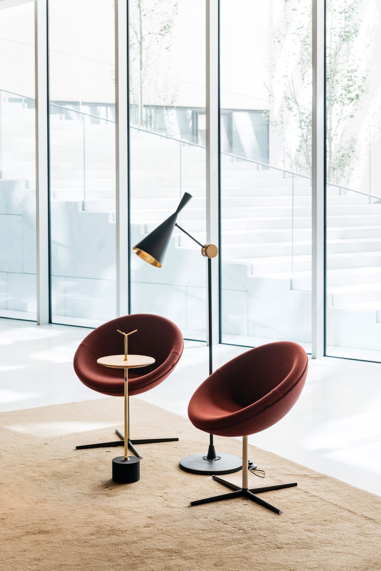 Two burgundy curved chairs with brass bases and a black articulating floor lamp in a bright reception area with floor-to-ceiling windows.