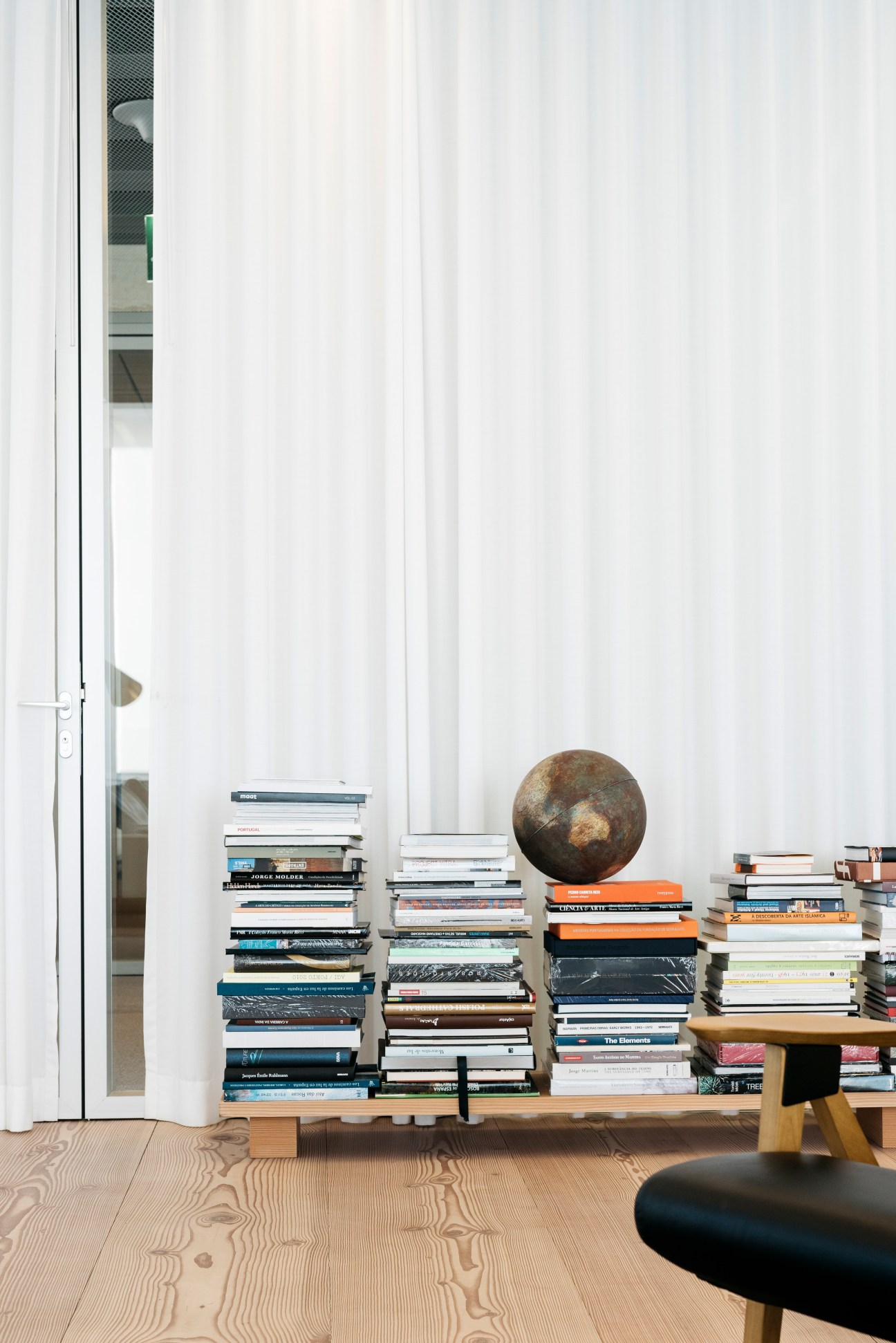 Stacked books with a bronze sphere decoration on a wooden shelf in an office space with white curtains.
