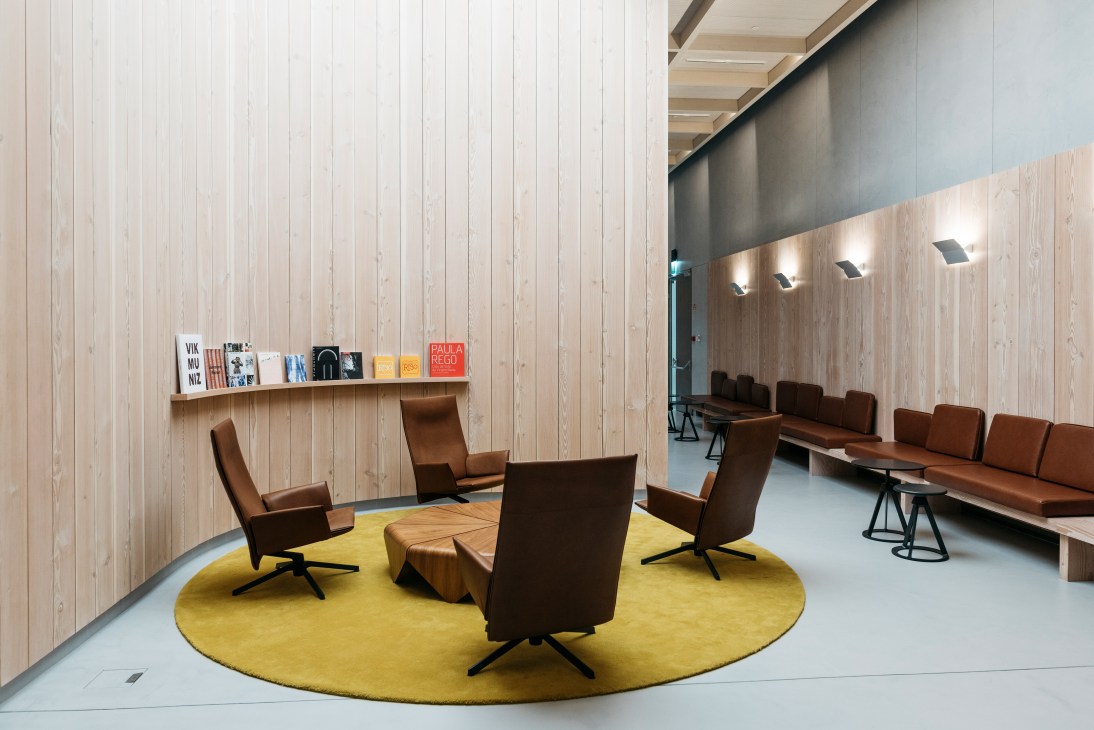 Modern meeting space with brown leather chairs arranged on a yellow circular rug, white wooden walls, and shelving with design books and publications.