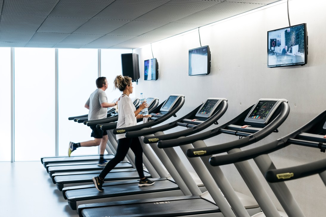 Two people running on treadmills in a modern gym with wall-mounted screens and white concrete architecture.