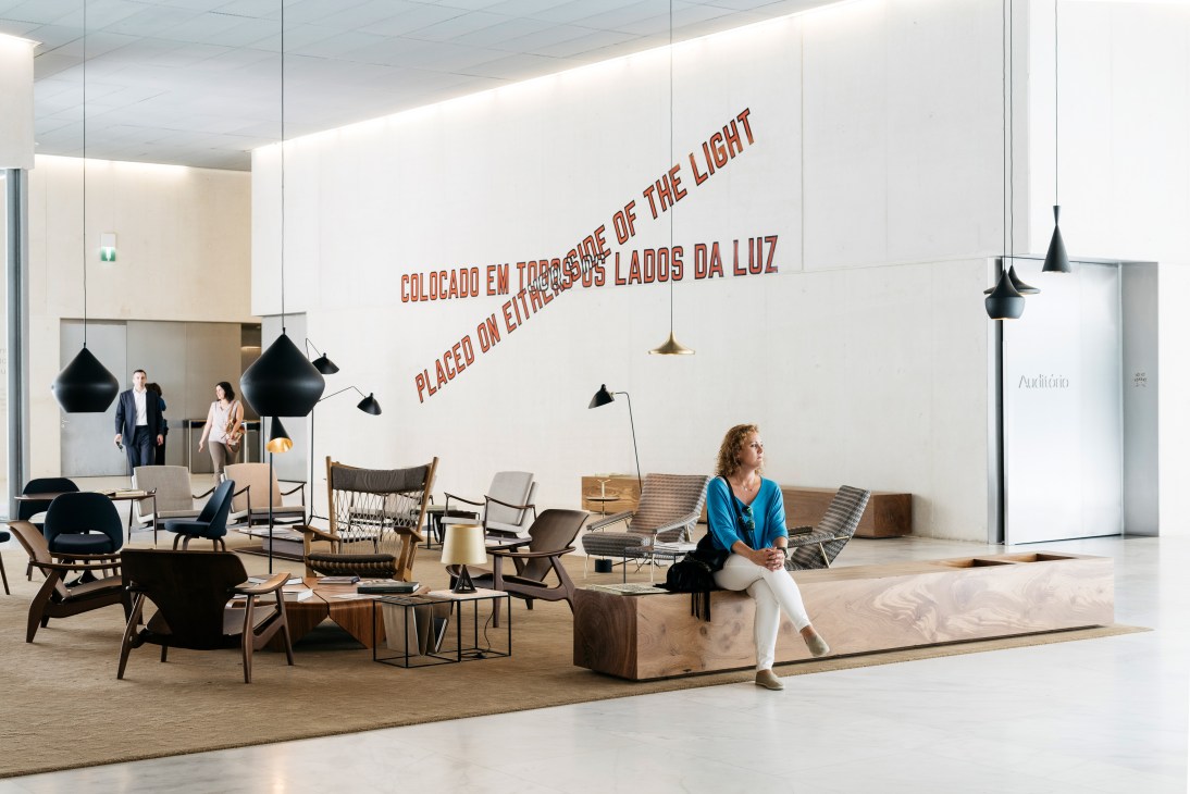 Modern reception area with high ceilings, black pendant lights, mid-century furniture, and a woman sitting on a wooden bench.