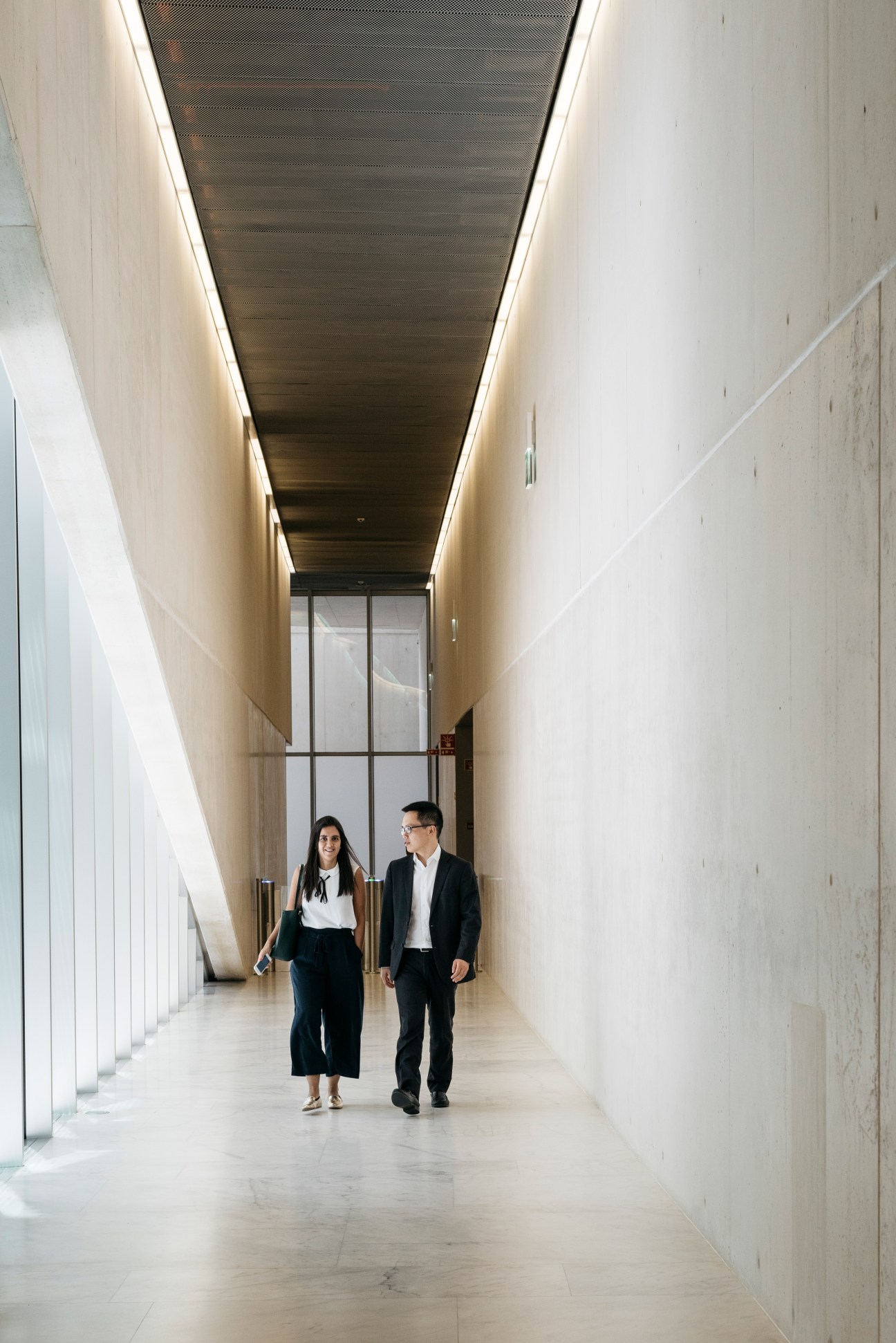 Two professionals walking through a minimalist corridor with six-metre high white concrete walls and linear ceiling lights.
