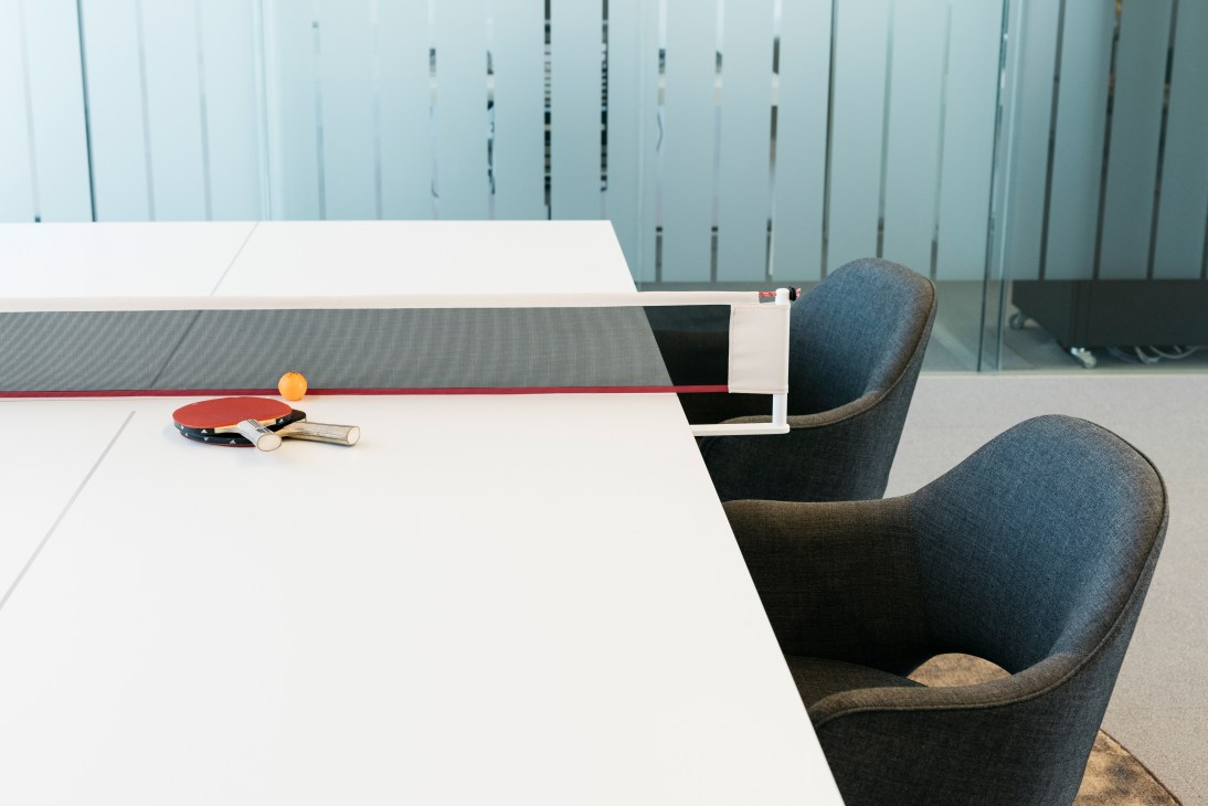 Ping-pong table with paddles and ball in a bright boardroom with blue upholstered chairs and glass walls.