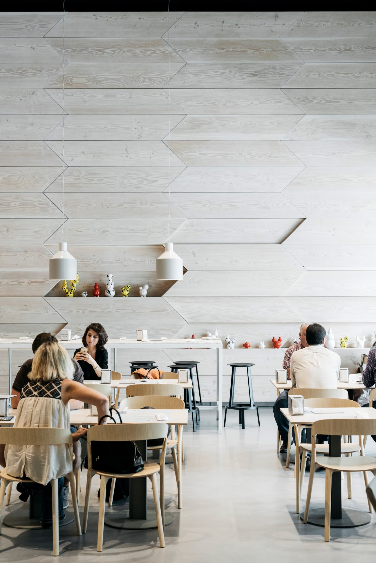 Modern canteen with light wood herringbone wall design, white ceramic vessels, and diners seated at round tables.