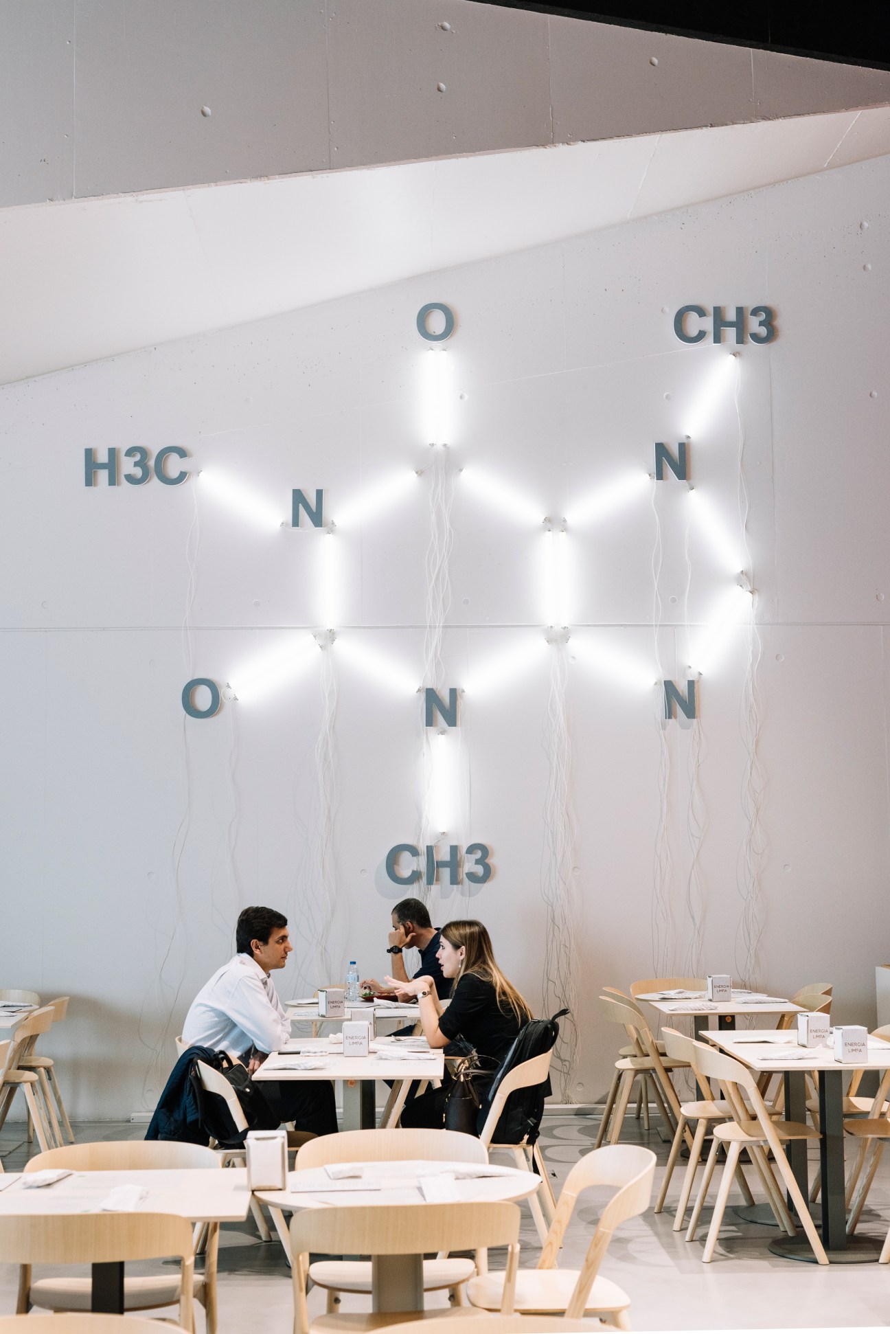 Canteen wall with illuminated neon chemical formula display and diners at wooden tables below.