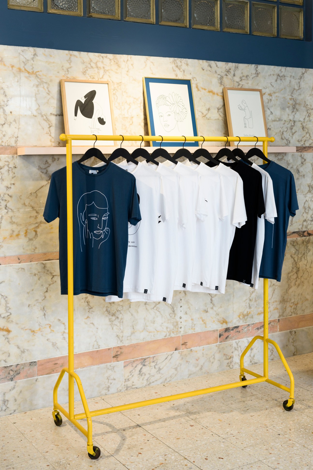 Yellow clothing rack with black hangers displaying navy, white and black t-shirts against marble wall with framed artwork above.