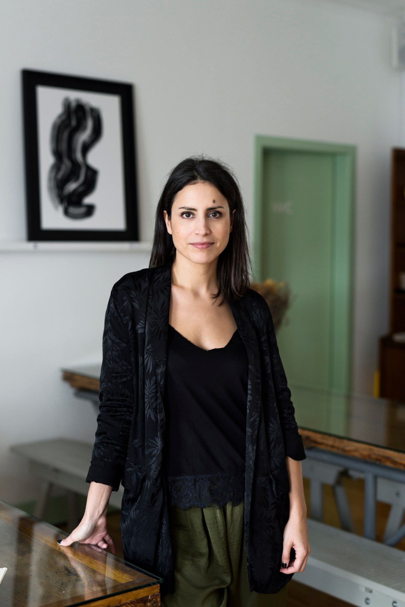 Ana Câmara, artistic director of Lapo, stands in the shop wearing a black cardigan over a black top with olive-green trousers, posing near framed artwork and a green door.