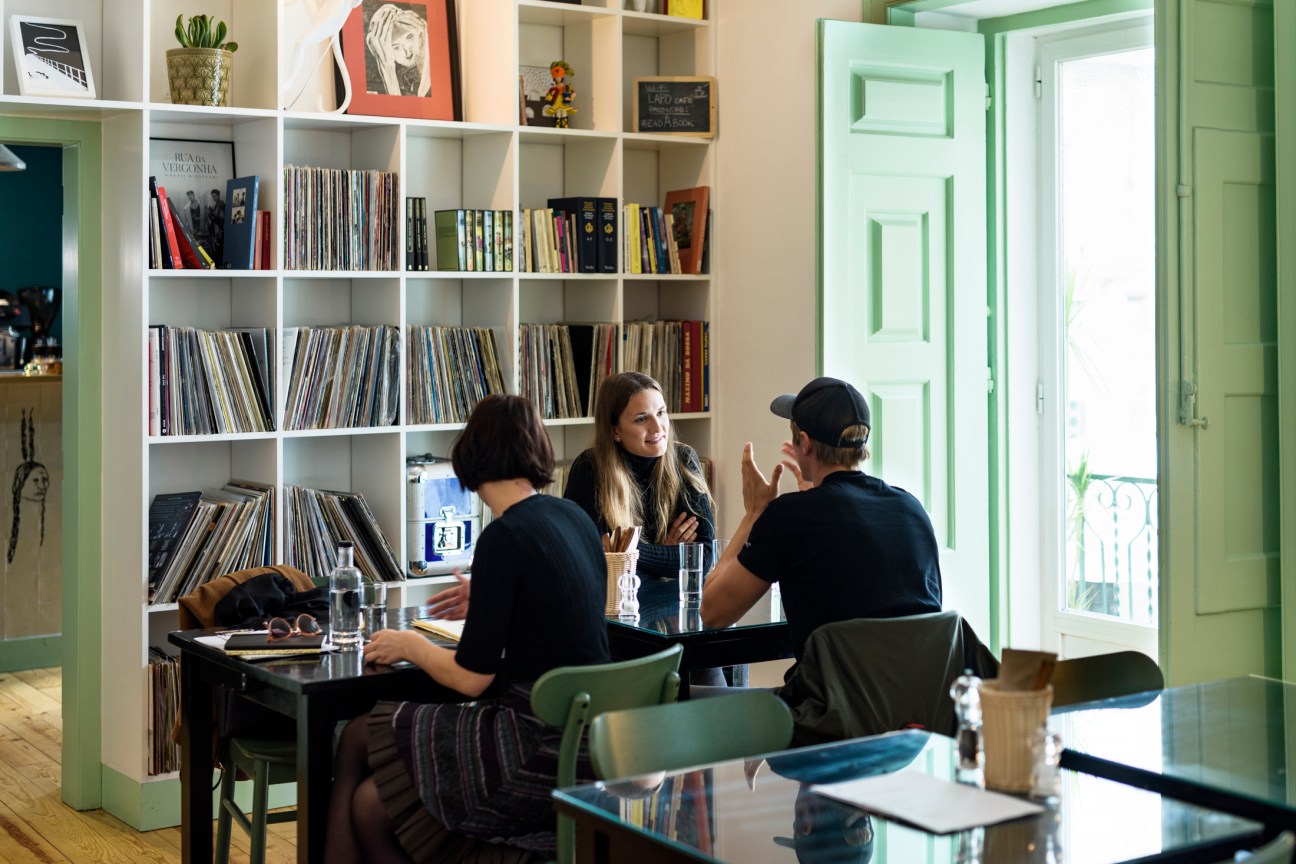 Three patrons sit at a table in a light-filled room with white shelving displaying books and vinyl records, with a green door visible in the background.