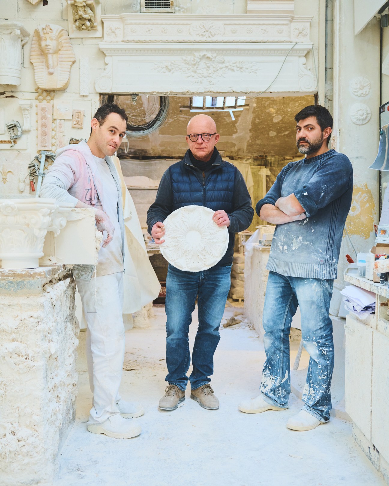 Three men stand in a plaster workshop holding a white circular cast piece, surrounded by decorative plaster elements on the walls.