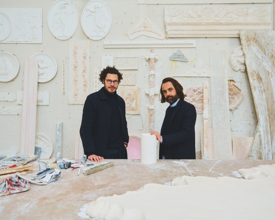 Florent Linker and Guillaume Garnier stand at a work table in a plasterworks studio surrounded by white plaster cast samples and decorative elements on the wall behind them.