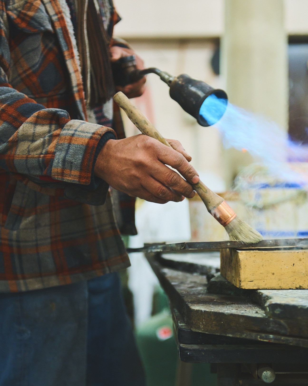 Artisan blow-torching bronze with a blue flame torch while holding a wooden tool.