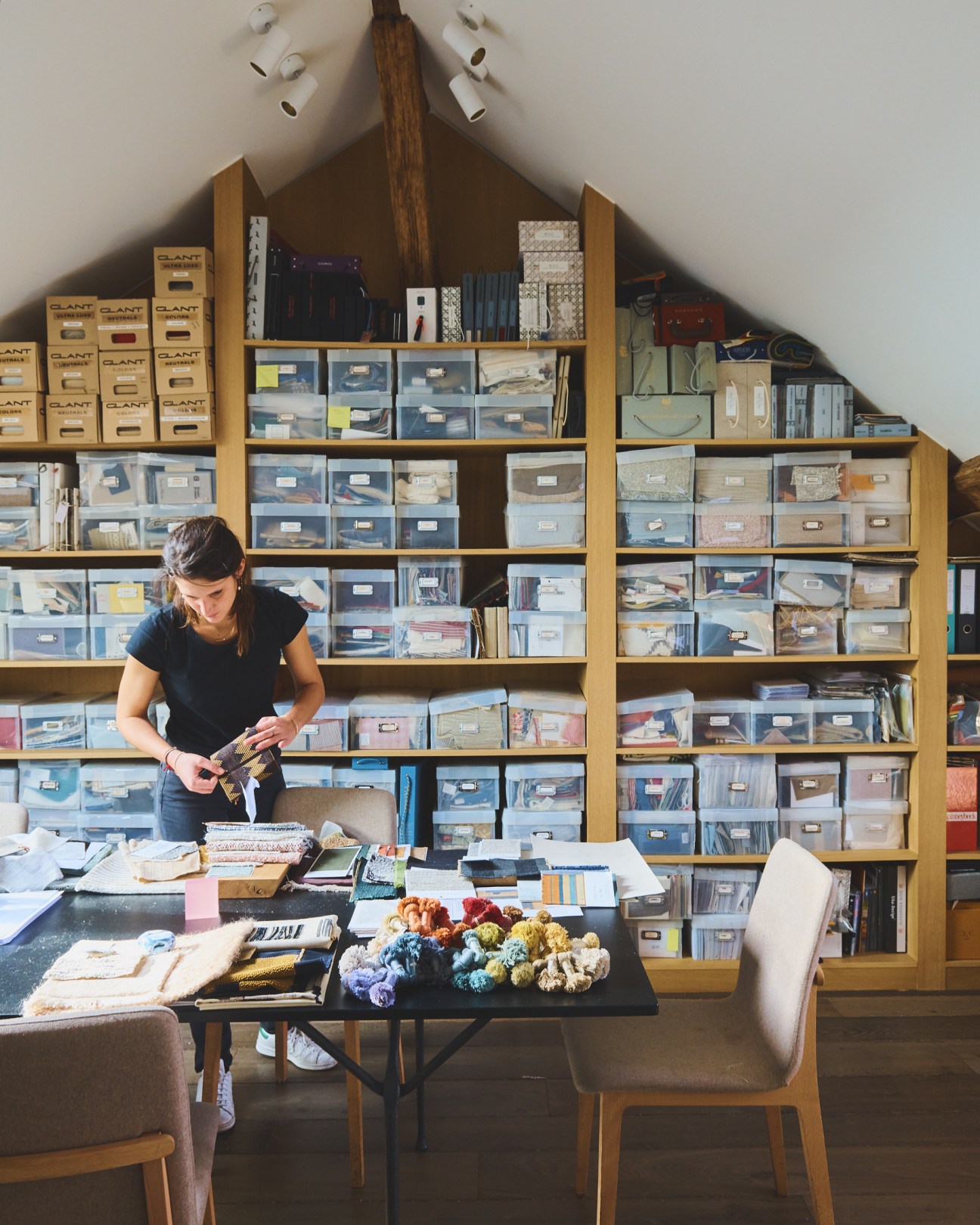 Woman working at a desk in a loft space with extensive wooden shelving displaying fabric and material samples.