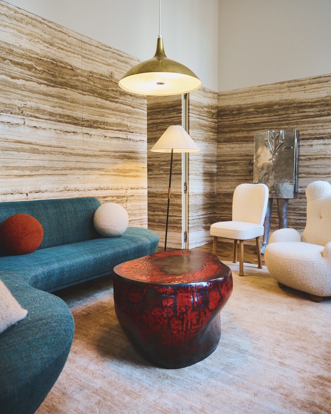 Modern interior with teal sofa, cream chairs, red-glazed side table, brass pendant light and travertine walls.
