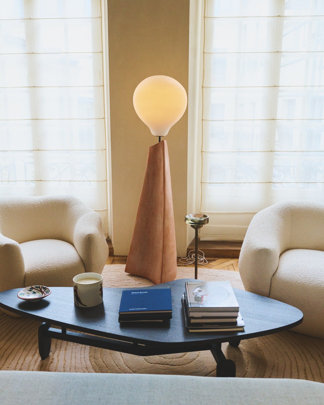 Modern interior space with cream armchairs, round table with books, and wooden cone-based floor lamp with glowing orb shade.