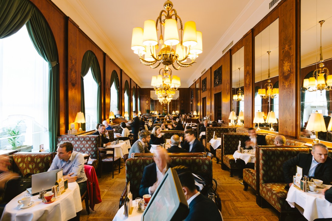 Elegant opulent coffeehouse with high ceilings, crystal chandeliers, wood paneling, and diners seated at tables.