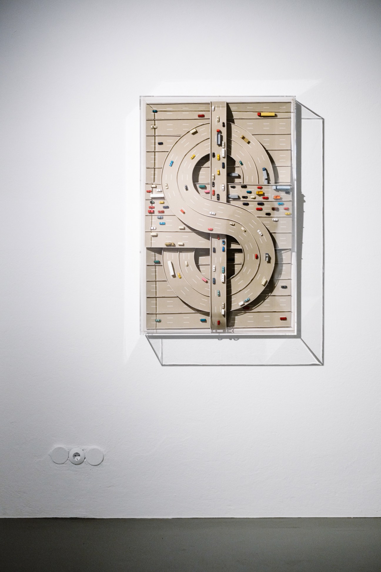 Overhead view of Thomas Bayrle's sculptural artwork featuring a dollar sign shape with curved roads and miniature vehicles on a wooden base displayed in a glass case.