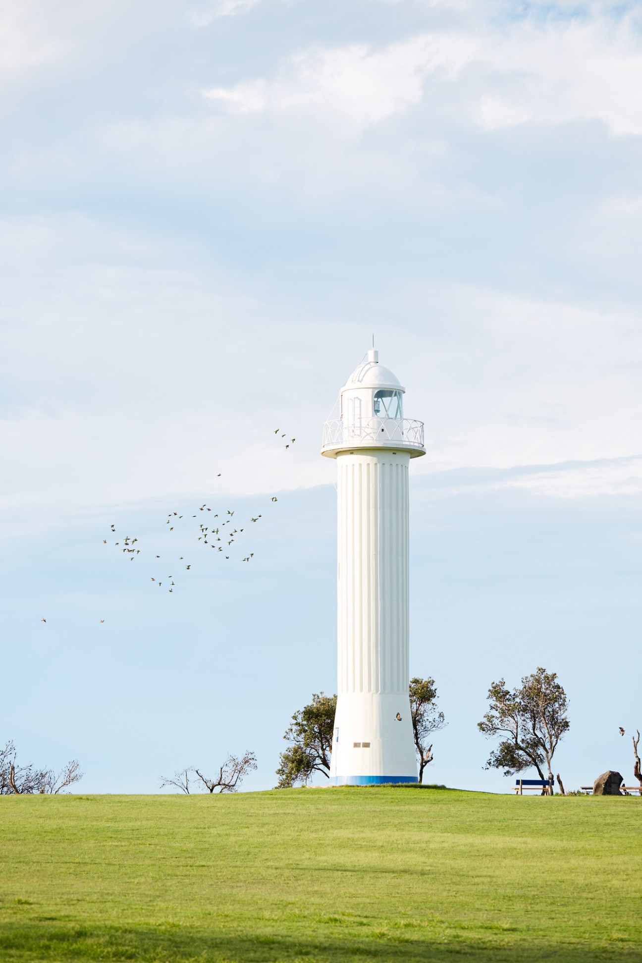 White lighthouse with dome top standing in a green field with birds flying nearby under a cloudy sky.
