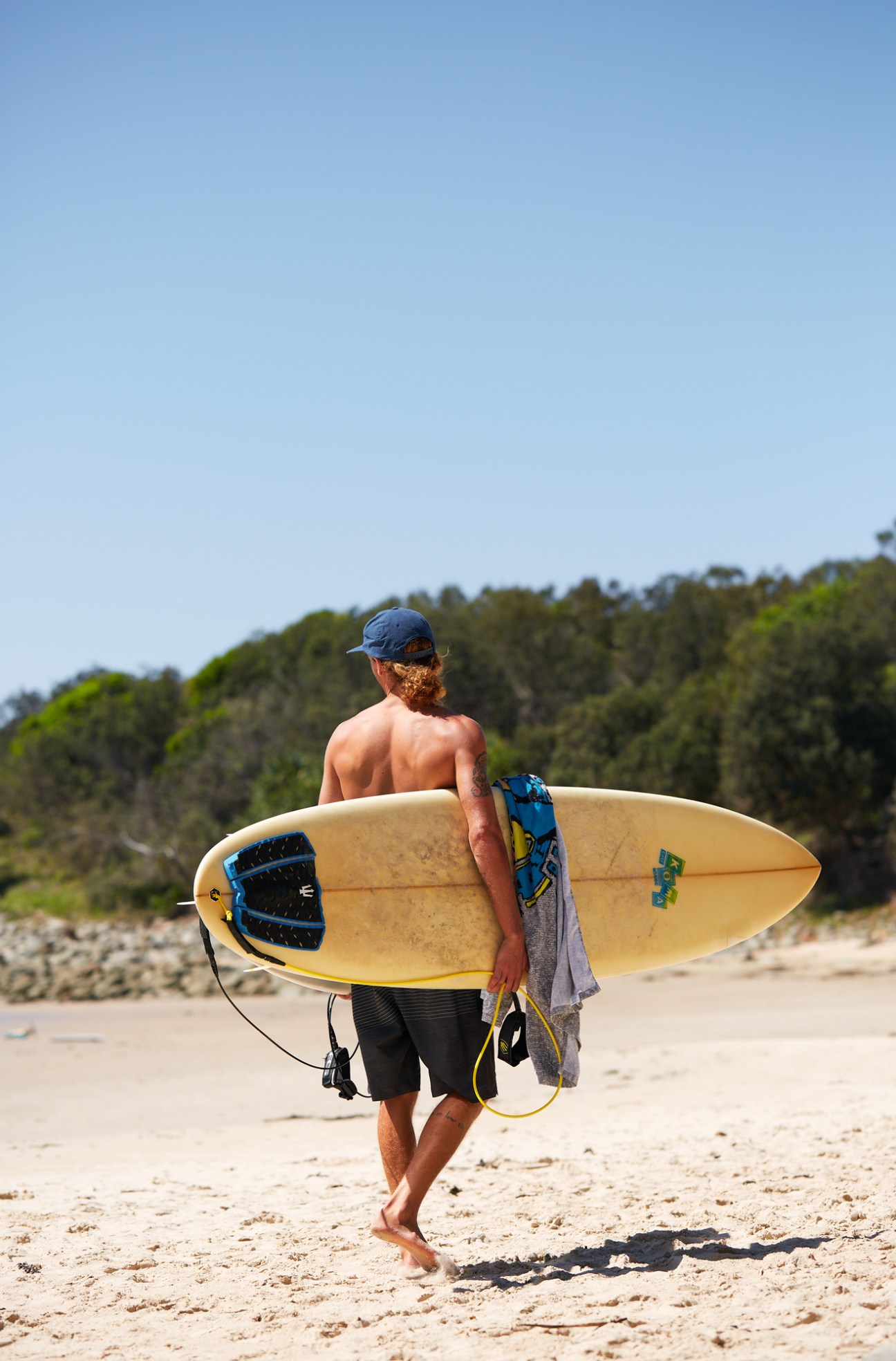 Shirtless surfer holding a yellow surfboard walks on sandy beach with forested headland in background.