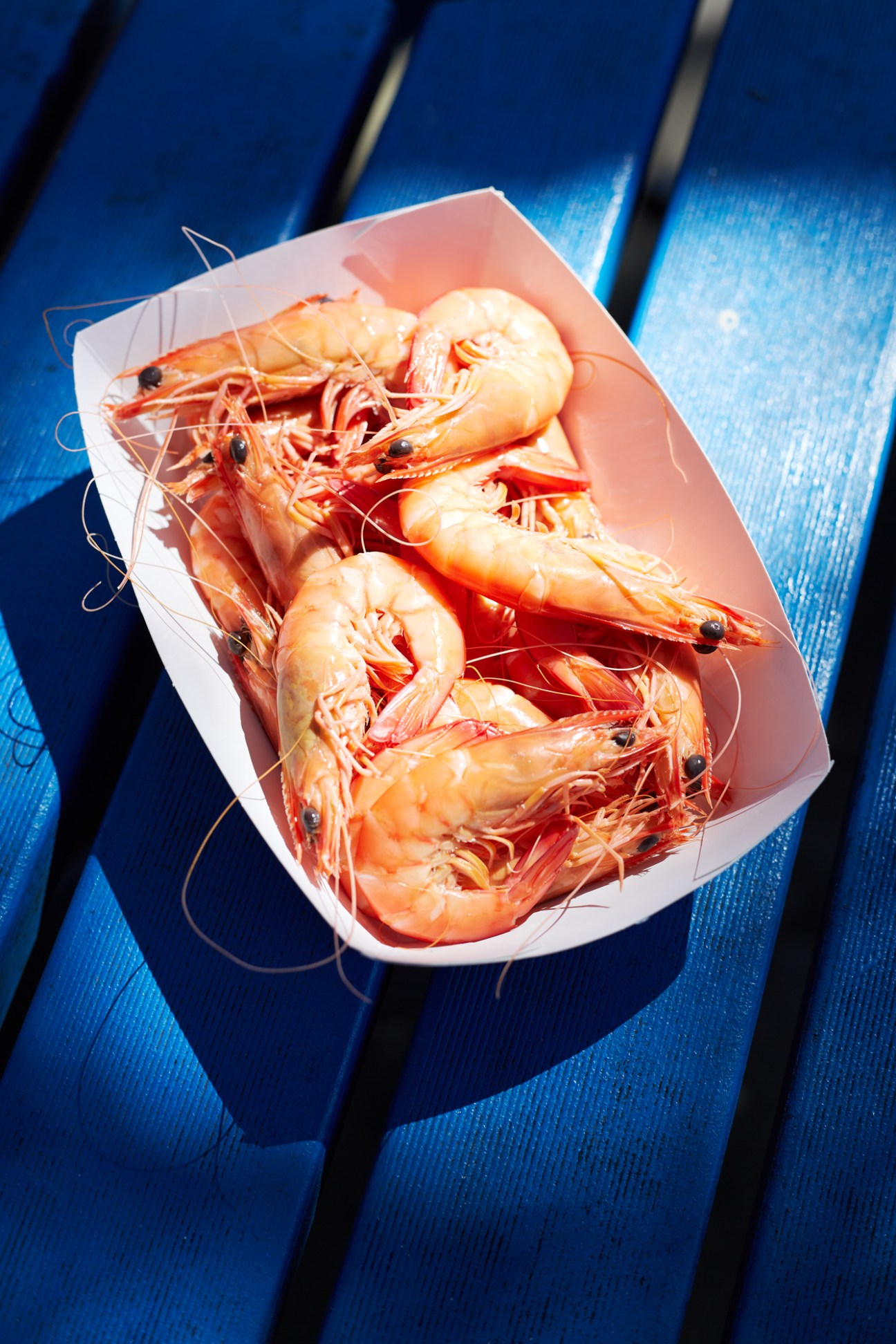 Fresh cooked prawns served in a white paper boat on a bright blue wooden surface.